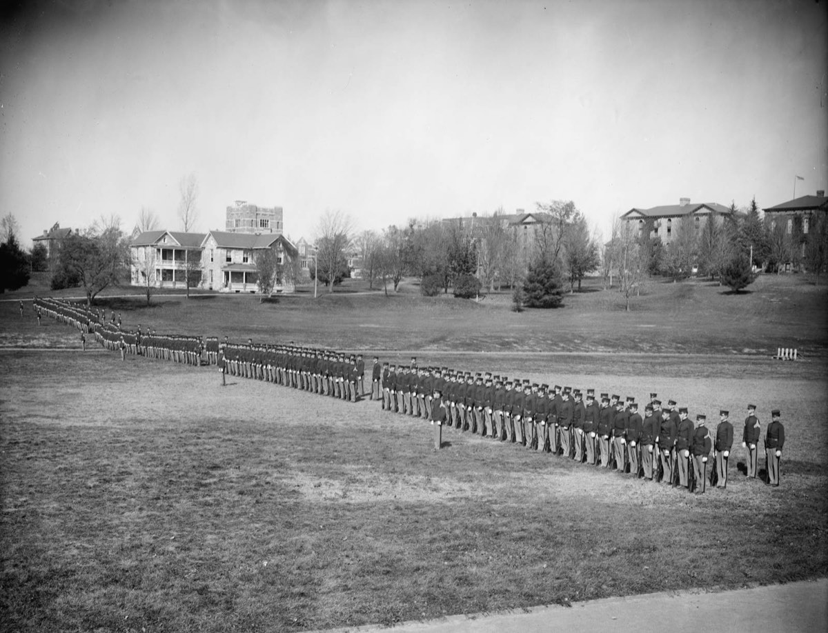 A long line of uniformed cadets standing in formation across a wide lawn, with large college buildings behind them